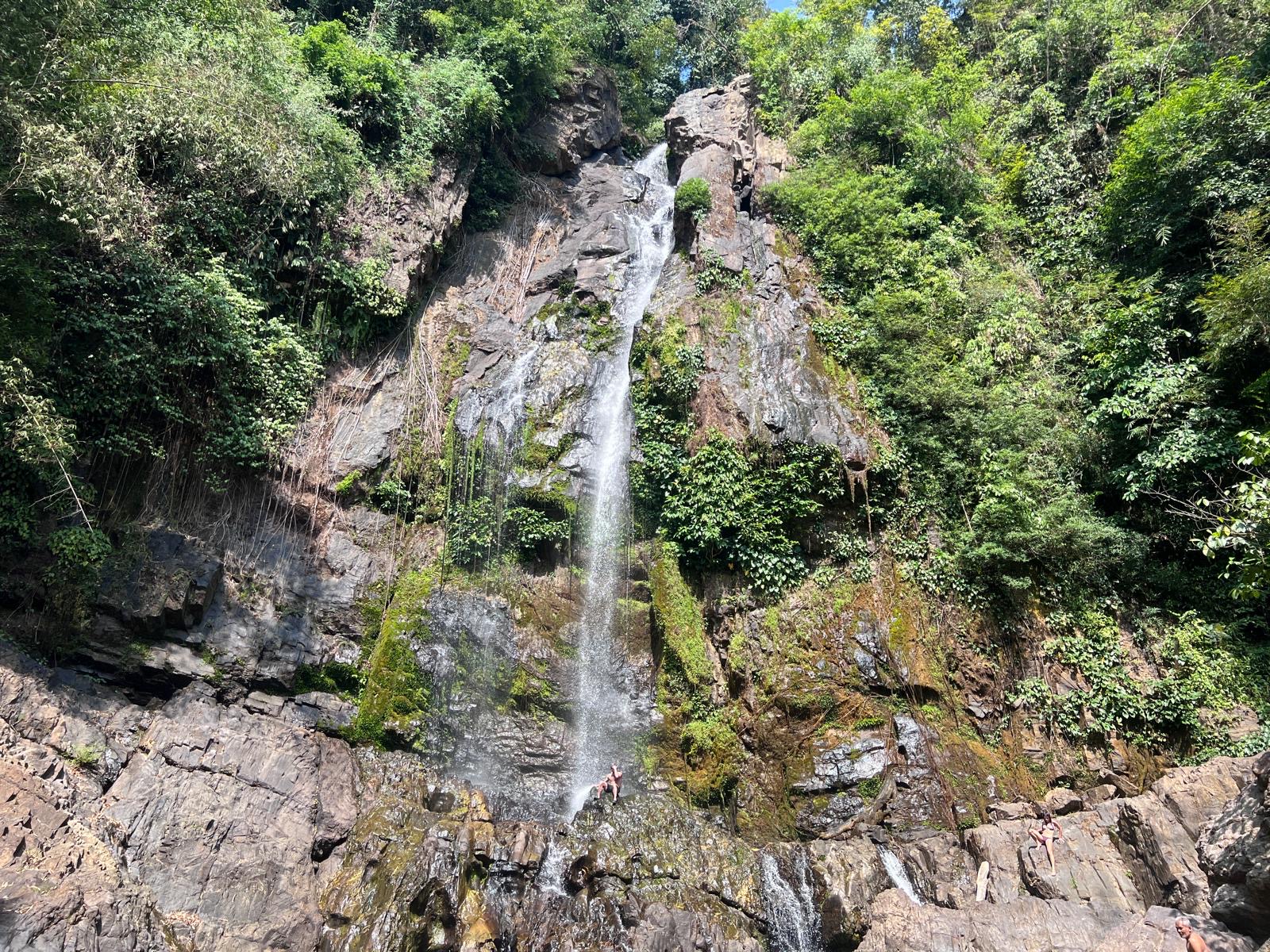 Een hoge, smalle waterval die over een donkere rotswand met weelderige begroeiing naar beneden stroomt.