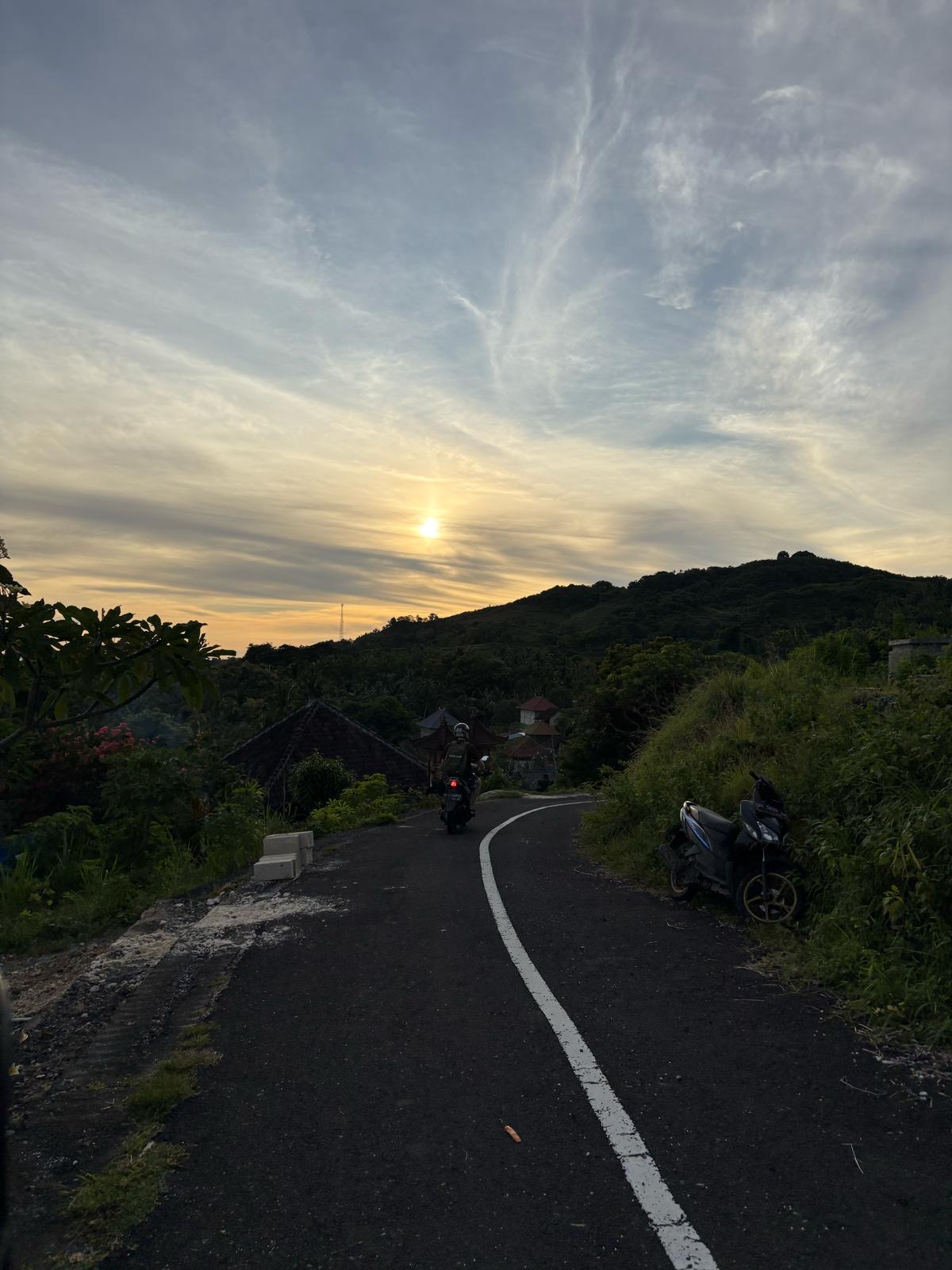 Een geasfalteerde weg op een heuvel met een rijdende scooter tijdens zonsondergang over het dal.