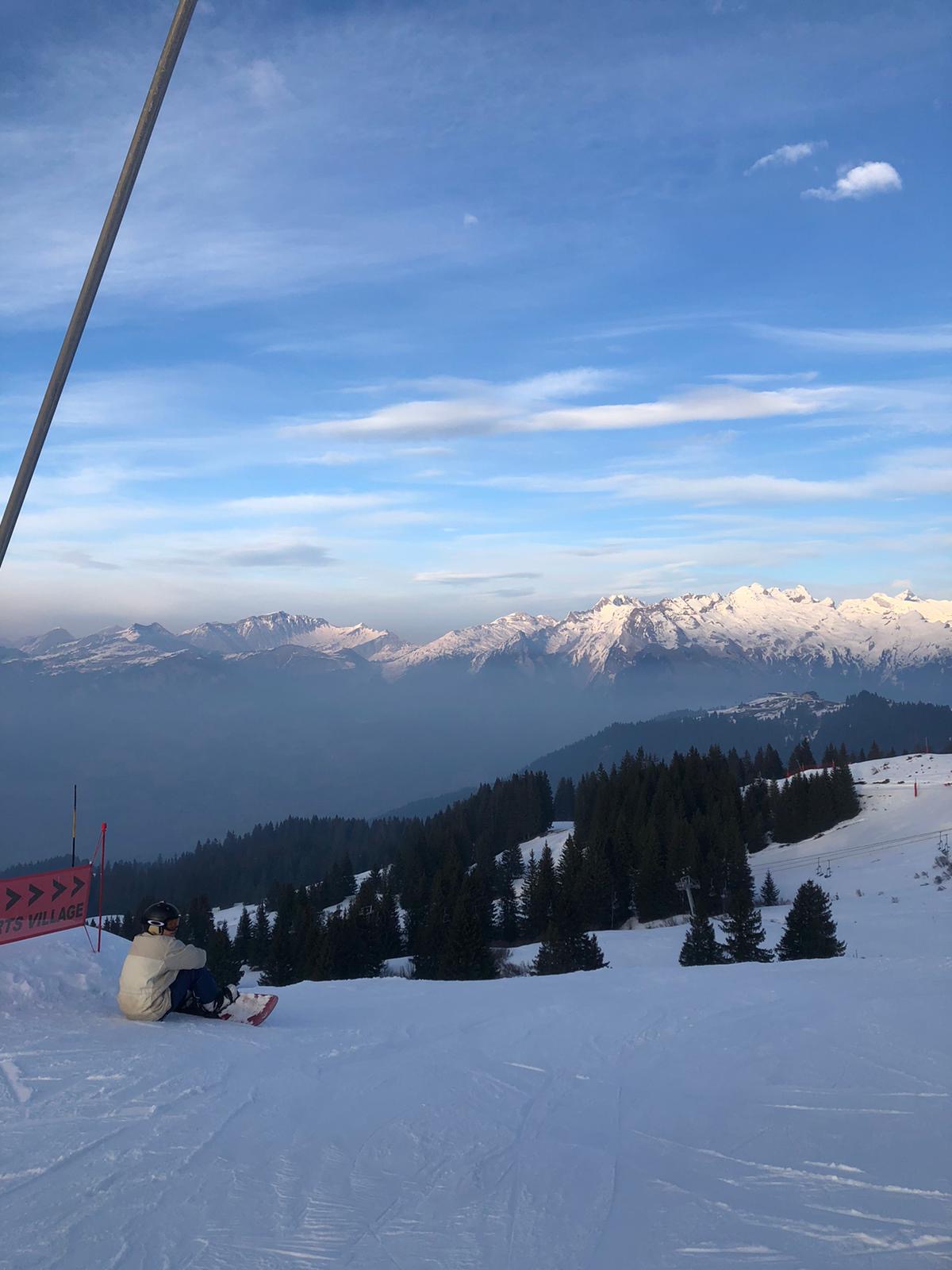 Berghelling met sneeuw en naaldbomen die uitkijkt over een zacht wolkendek en bergruggen in de verte.