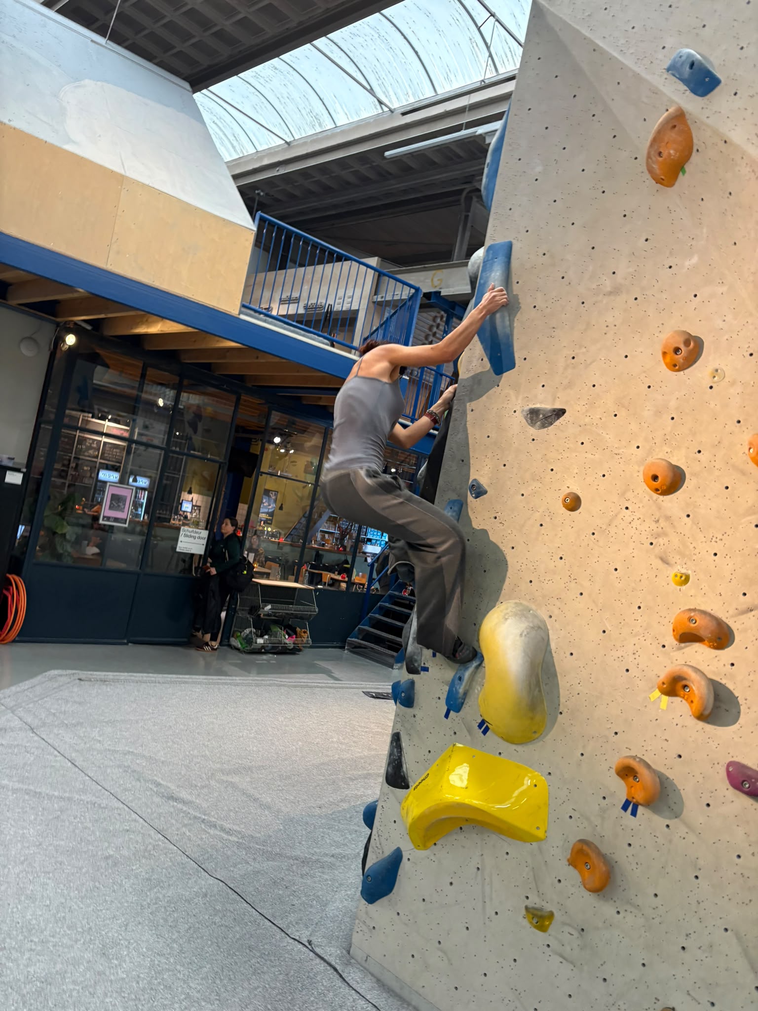 Zijaanzicht van Sophia in een grijze tanktop en losse broek die omhoog klimt op een indoor boulderwand. Sophia houdt zich vast aan een blauwe greep.