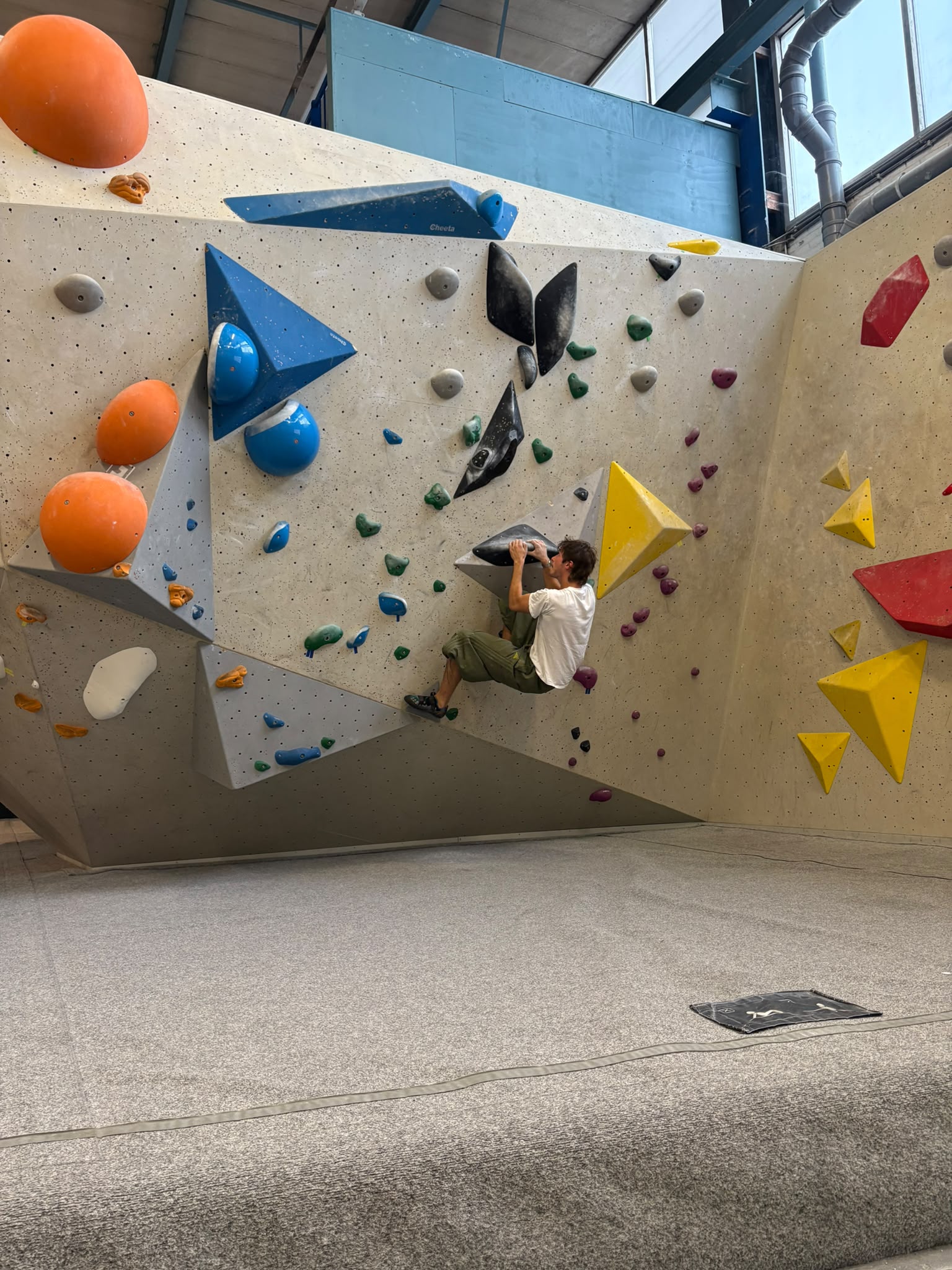 Steijn, in een wit T-shirt en groene broek hangt geconcentreerd aan een zwarte greep op een grote, hoekige boulderwand.