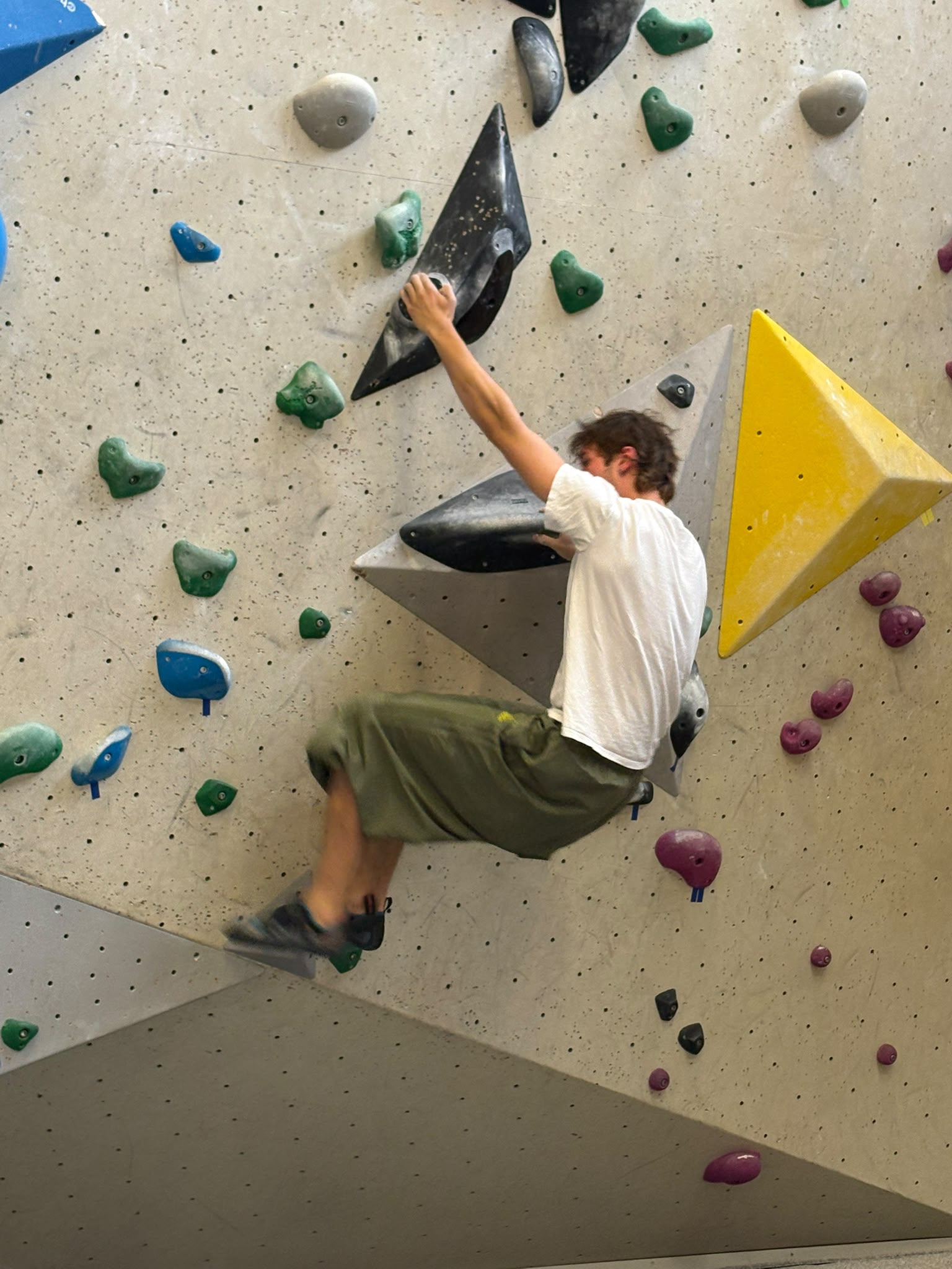 Steijn, in een wit T-shirt en groene broek hangt geconcentreerd aan een zwarte greep op een grote, hoekige boulderwand.