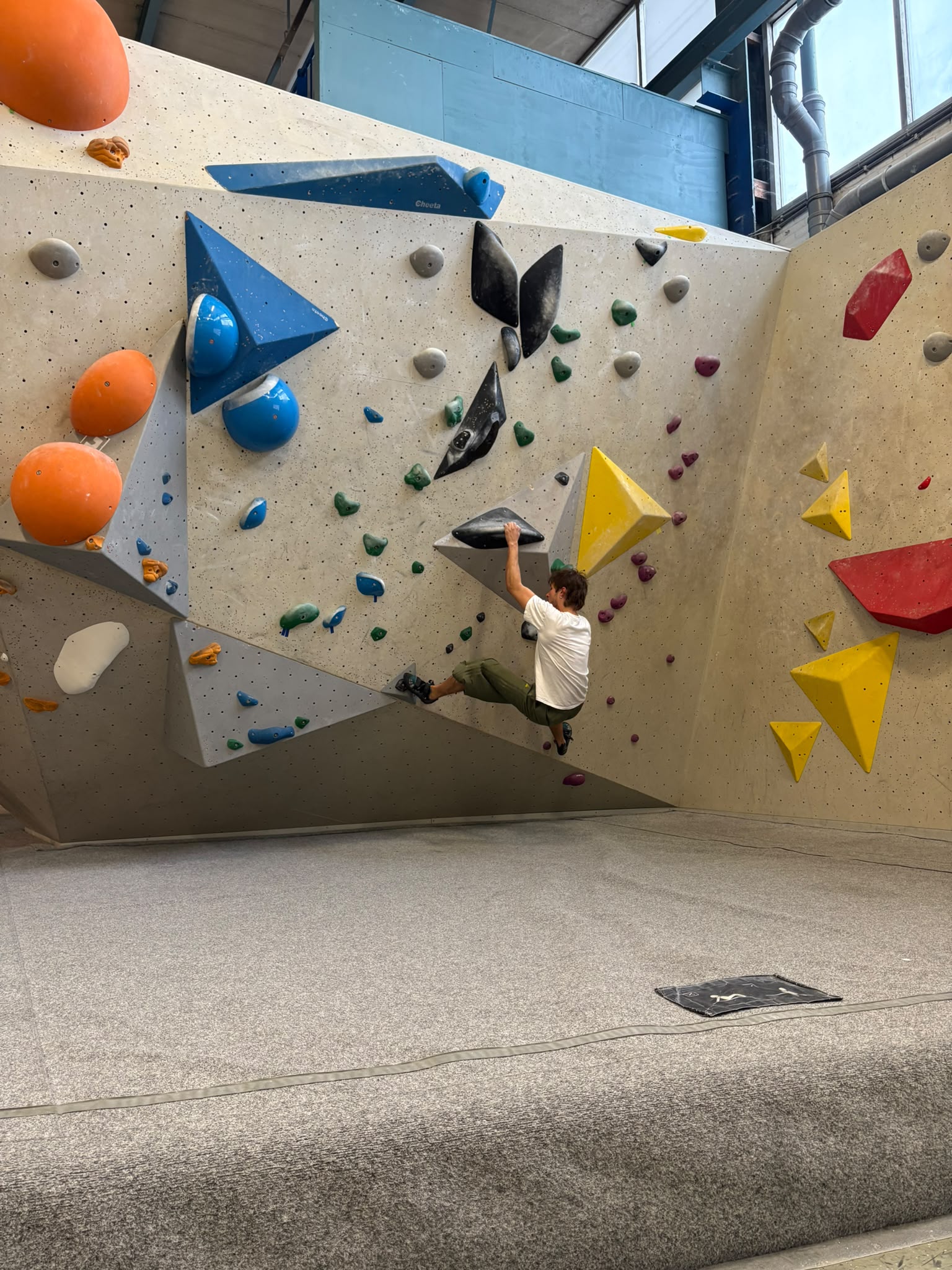 Steijn, in een wit T-shirt en groene broek hangt geconcentreerd aan een zwarte greep op een grote, hoekige boulderwand.