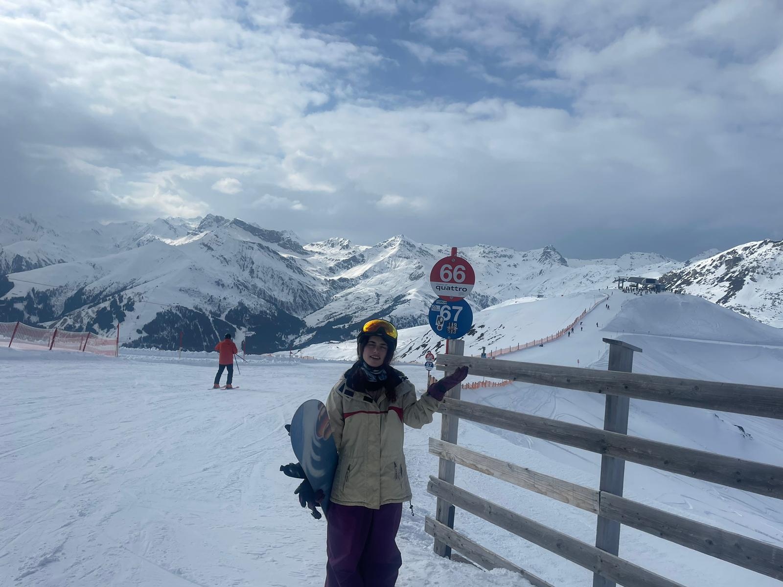 Een snowboarder in winterkleding met skibril en helm staat naast een houten hek op een besneeuwde piste met spitse bergtoppen op de achtergrond.