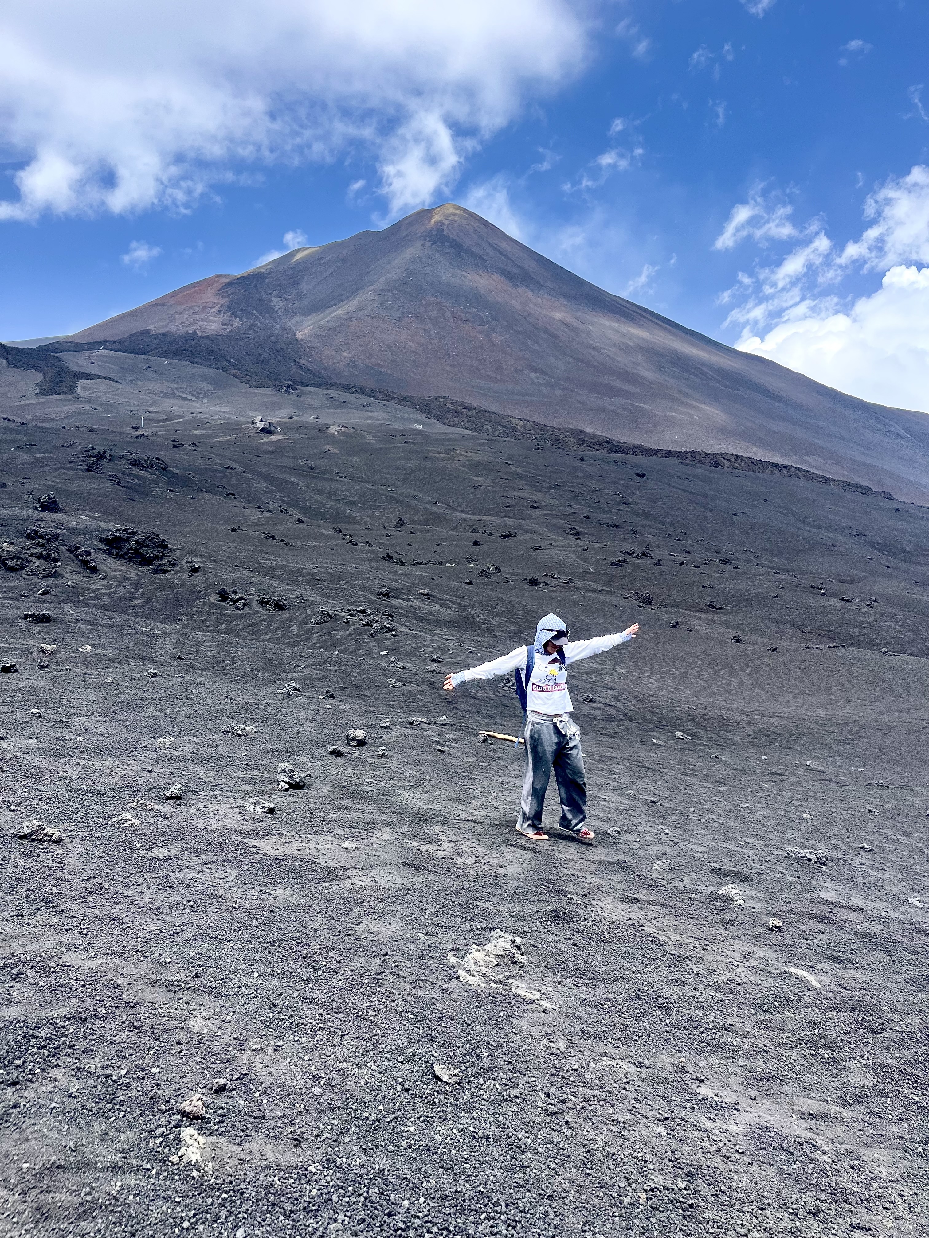 Een persoon in lichte kleding met open armen op een uitgestrekte, grijze vulkaanashelling met de top van de Etna op de achtergrond.